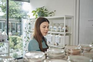 een wit vrouw winkelier cheques voorraad van natuurlijk biologisch producten in herbruikbaar glas potten Aan een plank Bij een bijvullen op te slaan, nul afval, plastic-vrij kruidenier winkel, en milieuvriendelijk, duurzame bedrijf. foto