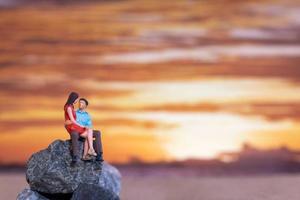 miniatuur mensen , paar zittend Aan een zee strand met zonsondergang achtergrond foto