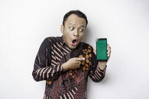 geschokt Aziatisch Mens vervelend batik overhemd en tonen groen scherm Aan zijn telefoon, geïsoleerd door wit achtergrond foto