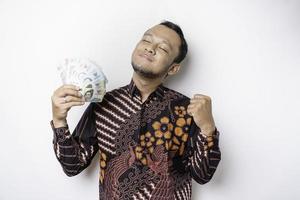 een gelukkig jong Aziatisch Mens is vervelend batik overhemd en Holding contant geld geld in Indonesisch roepia geïsoleerd door wit achtergrond foto