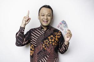 een gelukkig jong Aziatisch Mens is vervelend batik shirt, richten Bij de kopiëren ruimte Aan top van hem terwijl Holding geld in Indonesisch roepia geïsoleerd door wit achtergrond foto