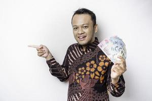 een gelukkig jong Aziatisch Mens is vervelend batik shirt, richten Bij de kopiëren ruimte naast hem terwijl Holding geld in Indonesisch roepia geïsoleerd door wit achtergrond foto