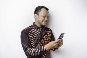 een portret van een gelukkig Aziatisch Mens vervelend batik overhemd en Holding zijn telefoon, geïsoleerd door wit achtergrond foto