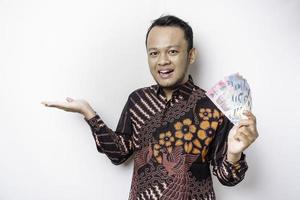 een gelukkig jong Aziatisch Mens is vervelend batik shirt, richten Bij de kopiëren ruimte naast hem terwijl Holding geld in Indonesisch roepia geïsoleerd door wit achtergrond foto