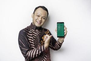 een portret van een glimlachen Aziatisch Mens vervelend een batik overhemd en tonen groen scherm Aan haar telefoon, geïsoleerd door wit achtergrond foto