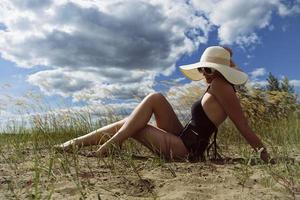 vrouw in een zwempak, hoed en zonnebril zonnebaadt in zomer Aan de oever van de rivier tussen de gras foto