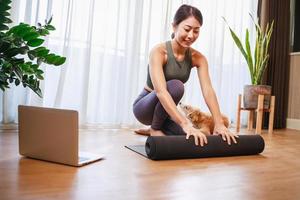 jong Aziatisch vrouw uitrollen rollen zwart yoga mat met haar hond Bij huis, nieuw normaal levensstijl concept foto