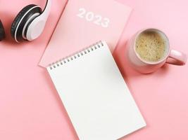 vlak leggen van blanco papier Notitie boek Aan roze dagboek of ontwerper 2023, hoofdtelefoons en roze kop van koffie Aan roze achtergrond met kopiëren ruimte. foto