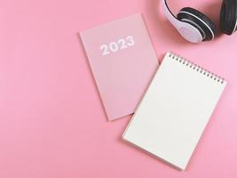 vlak leggen van blanco papier Notitie boek Aan roze dagboek of ontwerper 2023 en hoofdtelefoons Aan roze achtergrond met kopiëren ruimte. foto