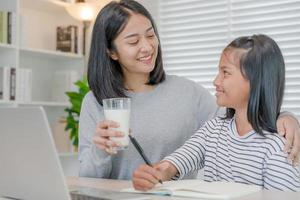 thuisschool Aziatisch jong weinig meisje leren, lezen en Doen huiswerk met soort moeder helpen, onderwijzen en aanmoedigen. mam voorbij gaan aan Aan een glas van melk naar dochter. meisje gelukkig naar studie en onderwijs samen met mam foto