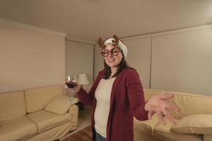 vrouw gekleed omhoog met rendier bril en Kerstmis hoed staand op zoek Bij de camera, roosteren met een glas van rood wijn in haar leven kamer Bij nacht foto