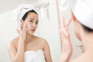 gelukkig jong Aziatisch vrouw toepassen gezicht lotions terwijl vervelend een handdoek en aanraken haar gezicht in badkamer foto