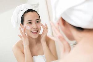 gelukkig jong Aziatisch vrouw toepassen gezicht lotions terwijl vervelend een handdoek en aanraken haar gezicht in badkamer foto