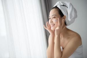 gelukkig jong Aziatisch vrouw toepassen gezicht lotions terwijl vervelend een handdoek en aanraken haar gezicht. dagelijks bedenken en huidsverzorging foto