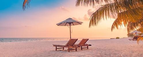 zomer strand landschap. luxe vakantie en vakantie concept, zomer reizen banner. panoramisch landschap van zonsondergangstrand, twee ligstoelenparaplu, palmblad, kleurrijke zonsonderganghemel voor uitzicht op het paradijselijke eiland foto