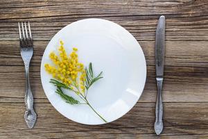 vork mes met bord en mimosa bloemen Aan houten tafel voor voorjaar Pasen vakantie diner. foto