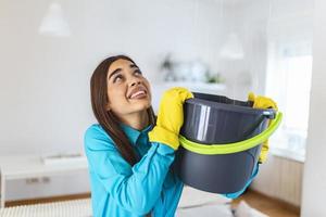 geschokt vrouw looks Bij de plafond terwijl verzamelen water welke lekken in de leven kamer Bij huis. bezorgd vrouw Holding emmer terwijl water druppels lekken van plafond in leven kamer foto