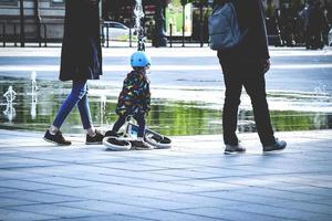 ouders met dochter wandelen Aan stad plein met fontein terwijl kind in helm is Holding een scooter in de buurt reusachtig water plas met groen bomen reflectie, mooi hoor familie moment foto