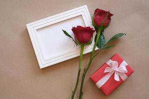 top visie van rood roos en rood geschenk doos houdende Aan een wit blanco foto kader geïsoleerd Aan bruin achtergrond. Valentijnsdag dag, bruiloft, verjaardag en speciaal gelegenheid concept. vlak leggen, top visie.