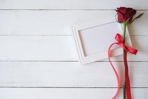 rood roos met rood lint Aan een wit blanco foto kader geïsoleerd Aan wit houten achtergrond. Valentijnsdag dag, bruiloft, verjaardag en speciaal gelegenheid concept. vlak leggen, top visie. kopiëren ruimte voor tekst.