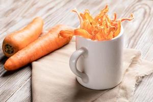 wit mok met lengte- wortel chips Aan beige kleding stof en twee geheel wortels Aan houten tafel. foto