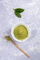 groen Japans matcha thee poeder in wit schaal, speciaal houten bamboe lepel en vers bladeren Aan grijs getextureerde achtergrond. foto