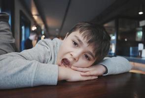 kind op zoek uit met verveeld gezicht, binnenshuis portret peuter- kind houdende hoofd naar beneden Aan hand- en geeuwen, jong jongen zittend in restaurant of cafe aan het wachten voor voedsel foto