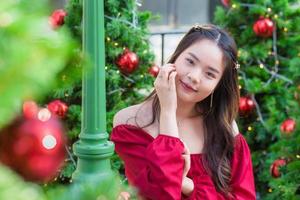 schattig Aziatisch vrouw in rood jurk staat glimlachen gelukkig op zoek Bij de camera in voorkant van de Kerstmis boom met bokeh net zo achtergrond in de thema van Kerstmis en nieuw jaar vieringen foto