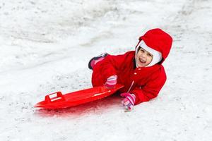 mooi glimlachen weinig meisje in haar ski pak glijden naar beneden een klein sneeuw gedekt heuvel met haar slee foto