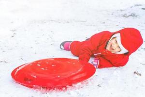 mooi glimlachen weinig meisje in haar ski pak glijden naar beneden een klein sneeuw gedekt heuvel met haar slee foto