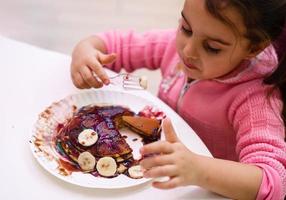 een weinig meisje eet pannekoeken met chocola en banaan foto