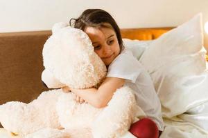 zoet weinig meisje is knuffelen een teddy beer op zoek Bij camera en glimlachen terwijl zittend Aan haar bed foto