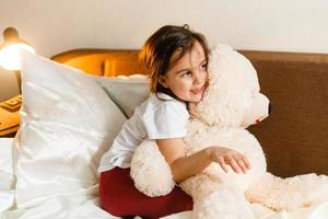 zoet weinig meisje is knuffelen een teddy beer op zoek Bij camera en glimlachen terwijl zittend Aan haar bed foto