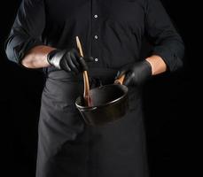mannetje koken in zwart uniform en latex handschoenen houdt een leeg ronde wijnoogst zwart gips ijzer pan foto