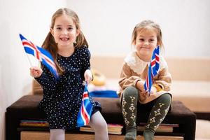 twee zussen zijn zittend Aan een bankstel Bij huis met IJslands vlaggen Aan handen. IJsland kinderen meisjes met vlag . foto