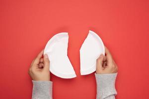 twee vrouw handen Holding helften van een plastic beschikbaar wit bord Aan een rood achtergrond foto