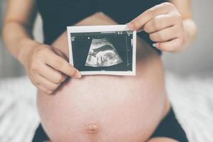 zwanger vrouw en haar man hand- las de buik Holding klein schoenen voor de ongeboren baby in de buik ontspannende Bij huis in slaapkamer. wacht geboorte datum ervan uitgaand een baby staan in de buitenshuis. foto