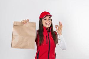 levering vrouw in rood uniform houden ambacht papier pakket met voedsel geïsoleerd Aan wit achtergrond, studio portret. vrouw werknemer in pet t-shirt afdrukken werken net zo koerier. onderhoud concept. foto
