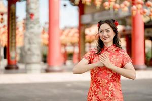Aziatisch vrouw vervelend traditioneel cheongsam qipao jurk met gebaar van felicitatie in Chinese boeddhistisch tempel. emotie glimlach. kettingen nieuw jaar concept foto