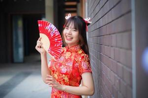 mooi Aziatisch jong vrouw in rood jurk staat glimlachen gelukkig op zoek Bij de camera houdt een ventilator tussen oud stad centrum in Chinese nieuw jaar thema. foto