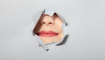vrouw tonen haar lippen speels in gescheurd papier gat, door doorbraak van grijs achtergrond. schoonheidsmiddelen reclame concept foto