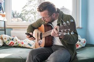 Mens met een akoestisch gitaar is aan het leren een nieuw melodie terwijl op zoek Bij aantekeningen Aan een smartphone. foto