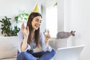 kopiëren ruimte schot van een vrolijk jong vrouw hebben een verjaardag viering evenement met een vriend over- een video telefoongesprek. ze is maken een feestelijk geroosterd brood met een glas van wit wijn naar laptop camera. foto