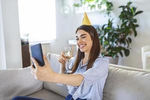 virtueel feest. gelukkig jong vrouw in hoed hebben video conferentie online vergadering met vrienden en familie, Holding glas van wijn, roosteren en vieren verjaardag, blijven Bij huis foto