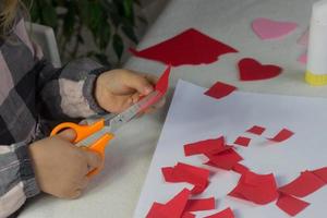 weinig meisje maken rood hart stoffen met papier en lijm Aan wit blad, valentijnsdag dag groet kaart, vakantie geschenk voor moeders dag, handen gemaakt kinderen heden, liefde symbool met papier, peuter werkzaamheid foto