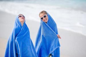 aanbiddelijk weinig meisjes verpakt in handdoek Bij tropisch strand na zwemmen in de zee. twee zussen spelen Aan de strand, strand handdoeken foto
