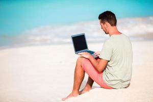 jong Mens zittend Aan zand met laptop Aan tropisch caraïben strand. Mens met computer en werken Aan de strand foto