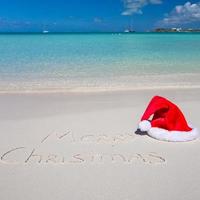 de kerstman hoed Aan wit zanderig strand en vrolijk Kerstmis geschreven in de zand foto