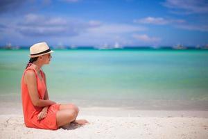 achterzijde visie van jong sexy vrouw in hoed zittend Aan wit zand strand foto