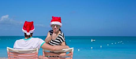 familie van twee in de kerstman hoeden zittend Aan strand stoel foto